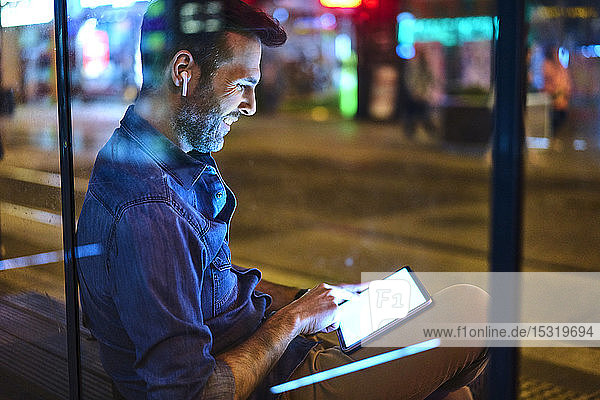 Mann mit Kopfhörern  der nachts an einem Bahnhof sitzt und sein digitales Tablett benutzt