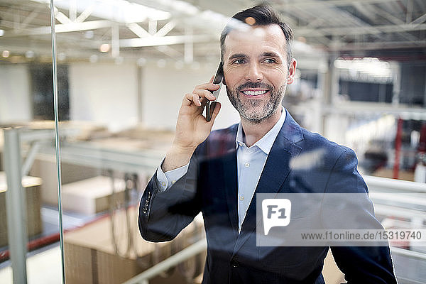 Porträt eines lächelnden Geschäftsmannes am Handy hinter Glasscheibe in einer Fabrik