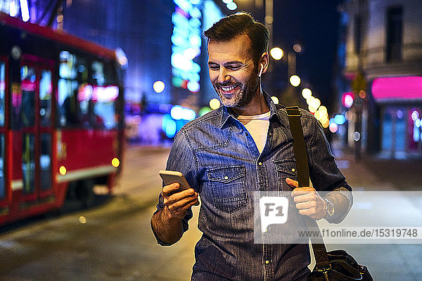 Smiling man with wireless headphones using smartphone while walking through the city at night