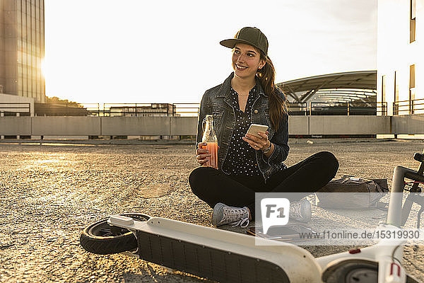 Lächelnde junge Frau mit Roller  Getränk und Handy sitzt bei Sonnenuntergang auf dem Parkdeck