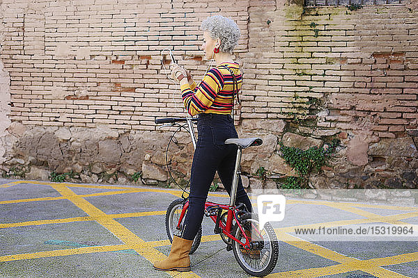 Modische reife Frau mit Fahrrad  die Ohrhörer und Smartphone benutzt