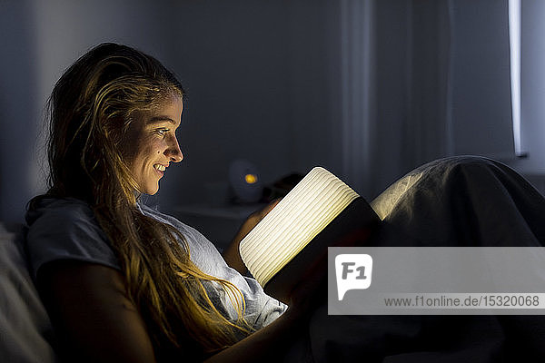 Smiling young woman reading illuminated book in bed at home