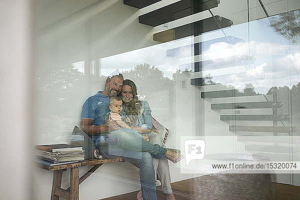 Glückliche dreiköpfige Familie sitzt zu Hause auf einer Bank hinter einer Fensterscheibe