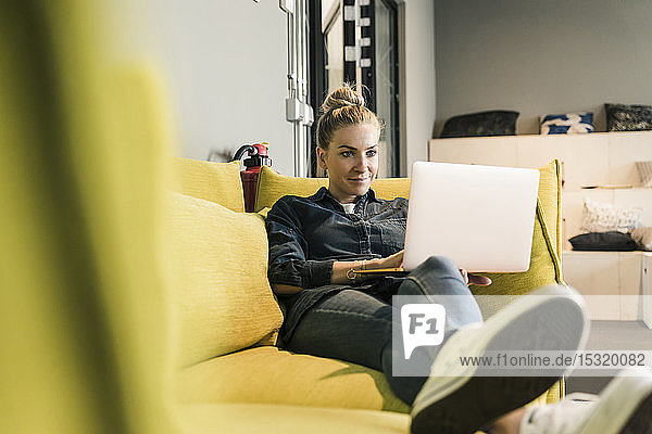 Casual businesswoman using laptop on couch in office lounge