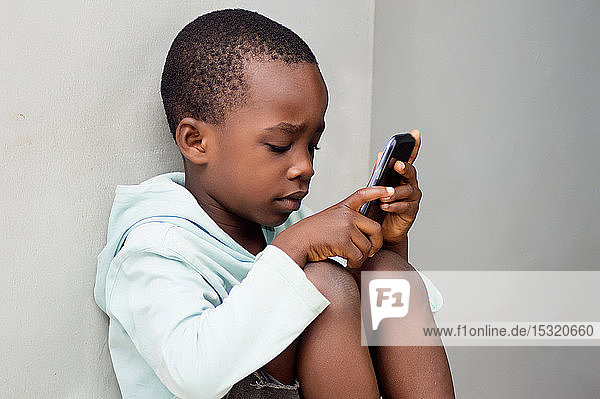 ein Kind  das an der Wand sitzt und neugierig mit einem Mobiltelefon hantiert.