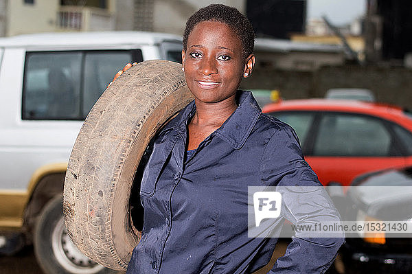 Junge Mechanikerin steht lächelnd mit einem Reifen auf der Schulter in ihrer Werkstatt.