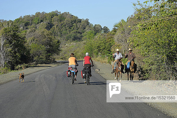 Cuba  eastern region  2 cyclo tourists are riding on an empty road where 2 men on horse are passing by with their dogs