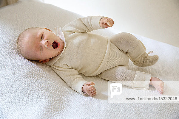 2 Monate altes Baby in weißer Jogginghose liegt auf dem Rücken in einem Bett.
