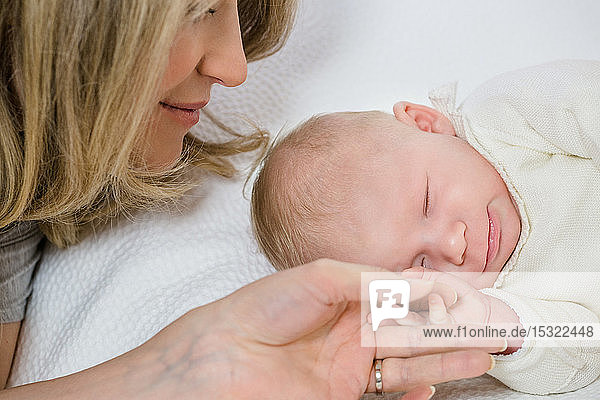 Nahaufnahme eines kleinen Säuglings in weißer Kleidung von 2 Monaten auf einem weißen Bett  der die Hand seiner Mutter hält.