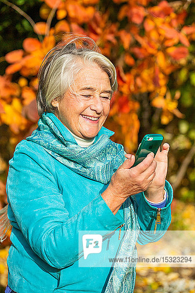Sonniges Porträt einer lächelnden hübschen älteren Frau mit seinem Telefon vor einem Baum mit herbstlichen Farben.