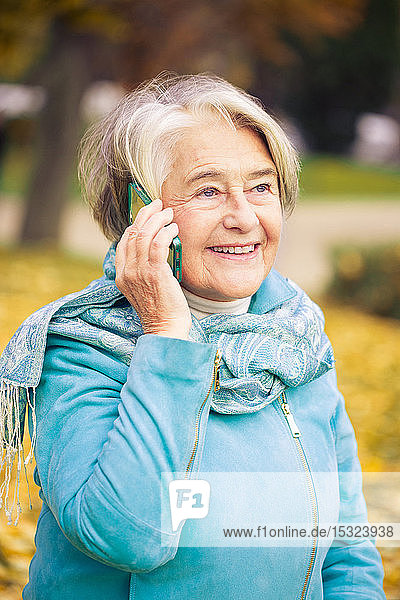 Sonniges Porträt einer lächelnden hübschen älteren Frau  die mit ihrem Telefon vor einem Baum mit herbstlichen Farben telefoniert.