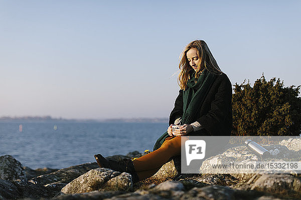 Frau mit Becher auf einem Felsen am Meer sitzend