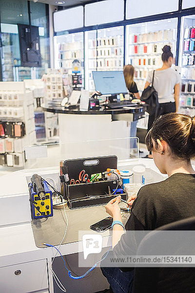Young female employee repairing mobile phone while customers standing in store.