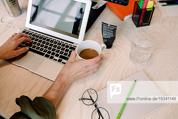 Schrägansicht einer Geschäftsfrau  die eine Kaffeetasse hält  während sie im Büro am Laptop arbeitet