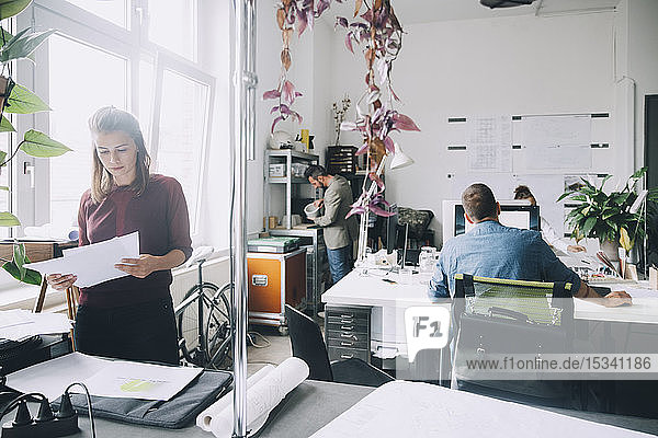 Creative businesswoman examining papers while colleagues working in background at office