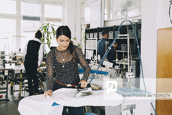 Female entrepreneur ironing white fabric while colleagues working in background at creative office