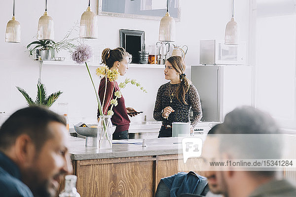 Kreative Unternehmerinnen diskutieren auf der Kücheninsel  während Kollegen im Büro im Vordergrund stehen