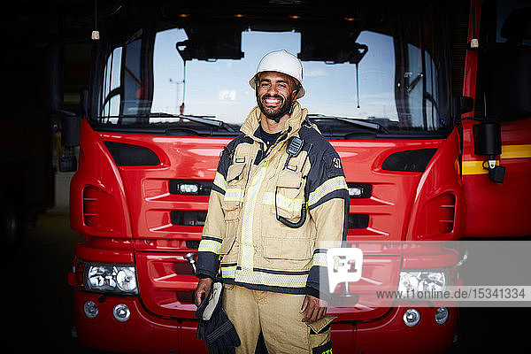 Porträt eines lächelnden Feuerwehrmannes in Uniform  der in der Feuerwache vor dem Feuerwehrauto steht