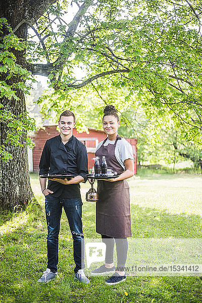 Full length portrait of smiling confident young male and female wait staff standing on grass at outdoor cafe