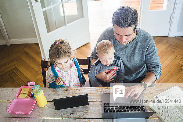 Hochwinkelansicht eines Vaters  der am Laptop arbeitet  während er mit dem süßen Sohn von der Tochter zusammensitzt und ein digitales Tablett am Tisch benutzt