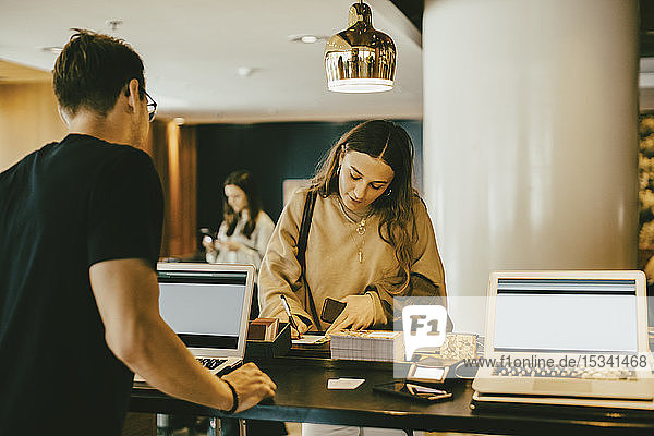 Man looking at woman signing at reception desk in hotel lobby
