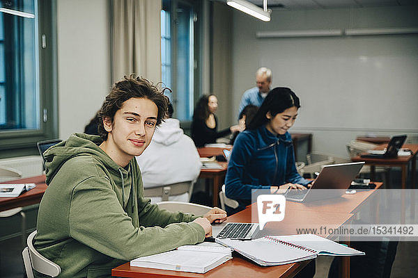 Porträt eines lächelnden Teenagers mit Laptop am Schreibtisch mit Klassenkameraden  die im Klassenzimmer lernen
