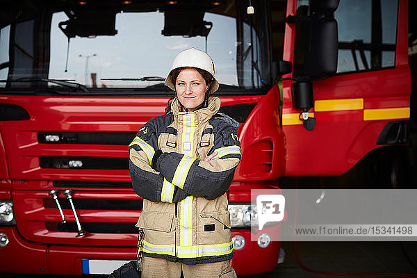 Porträt einer selbstbewussten Feuerwehrfrau  die mit verschränkten Armen gegen ein Feuerwehrauto auf der Feuerwache steht