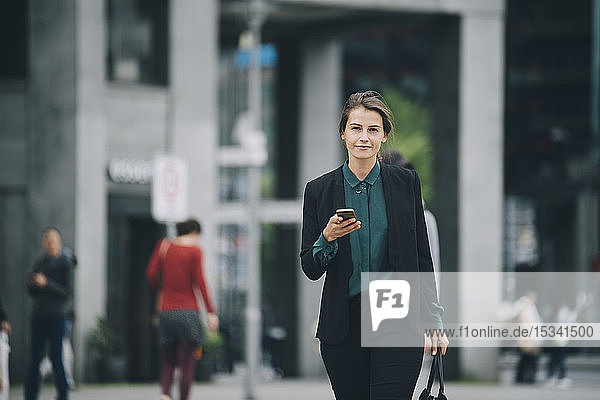 Porträt einer selbstbewussten Geschäftsfrau  die ein Smartphone in der Hand hält  während sie in der Stadt auf der Straße geht