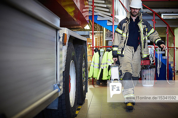 Full length of firefighter holding fire hose while walking by fire engine at station