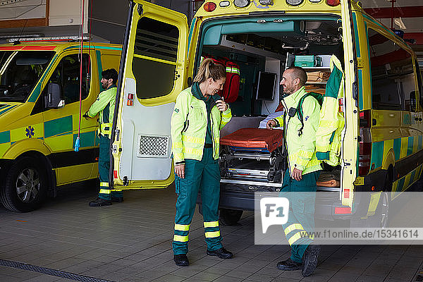 Lächelnder männlicher Sanitäter  der eine Mitarbeiterin ansieht  die mit einem Walkie-Talkie spricht  während er auf dem Parkplatz beim Krankenwagen steht