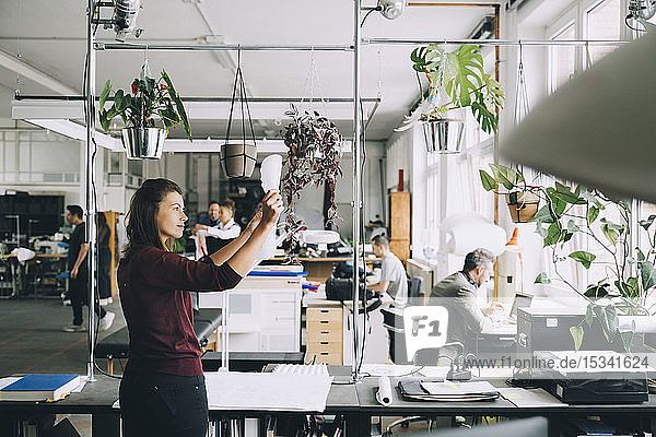 Mittlere erwachsene Geschäftsfrau prüft Papier  während Kollegen im Hintergrund im Kreativbüro arbeiten