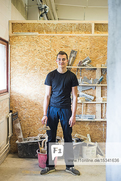 Porträt eines selbstbewussten jungen männlichen Auszubildenden in voller Länge  der mit Ausrüstung in der Werkstatt steht