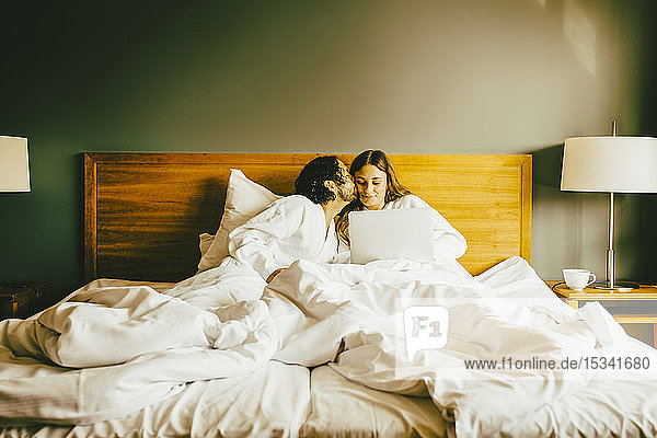 Mann küsst im Hotelzimmer seine am Laptop arbeitende Freundin im Bett