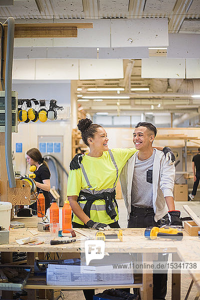 Cheerful female trainee standing with arm around male coworker at workbench