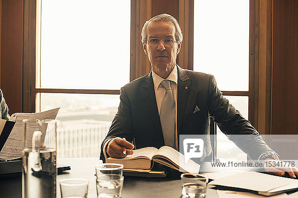 Portrait of confident mature lawyer with book at conference table in office