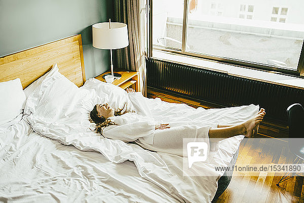 Lächelnde Frau im Bademantel amüsiert sich im Hotelzimmer im Bett
