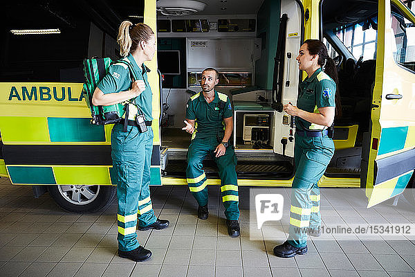Männlicher Rettungssanitäter sitzt im Krankenwagen  während er sich auf dem Parkplatz mit Kollegen unterhält