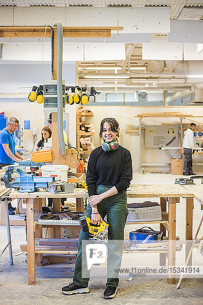 Full length portrait of smiling young female trainee holding power tool while standing in workshop