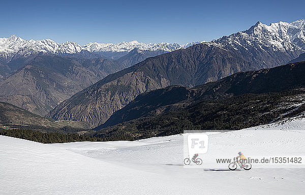 Mountain bikers descend a snow covered slope in the Himalayas with views of the Langtang range in the distance  Nepal  Asia