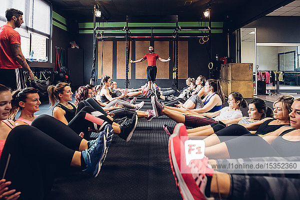 Große Gruppe von Frauen trainiert in Turnhalle mit männlichen Trainern  die mit angehobenen Beinen auf dem Boden sitzen