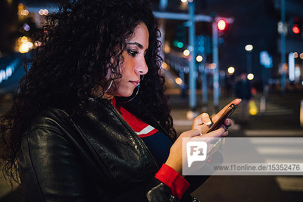 Mittelgroße erwachsene Frau mit langen lockigen Haaren  die nachts auf der Straße in der Stadt ein Smartphone betrachtet