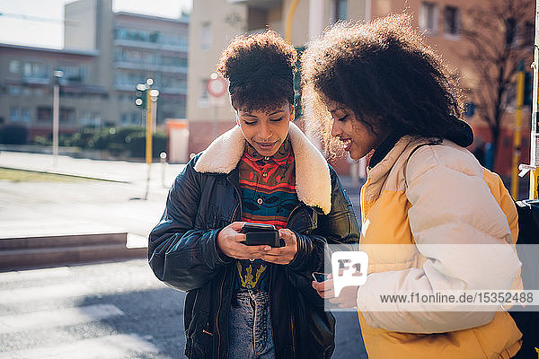 Two cool young women looking at smartphone on urban sidewalk