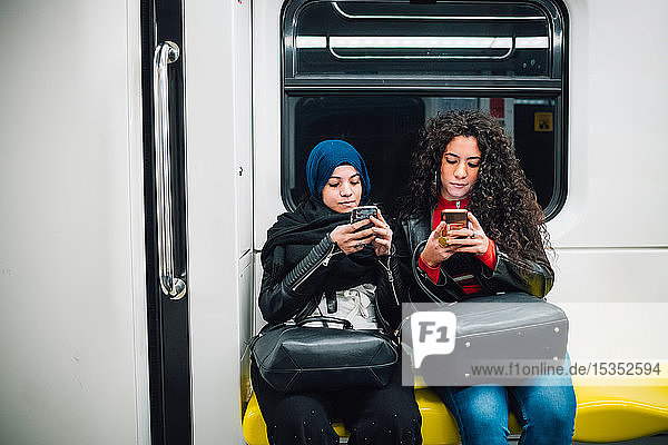 Junge Frau im Hidschab und Freundin sitzen in der U-Bahn und schauen sich Smartphones an