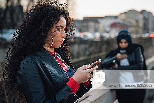 Mittelgroße erwachsene Frau mit langen lockigen Haaren betrachtet Smartphone am Stadtkanal