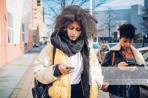 Zwei coole junge Frauen  die auf dem städtischen Bürgersteig spazieren gehen und sich Smartphones anschauen