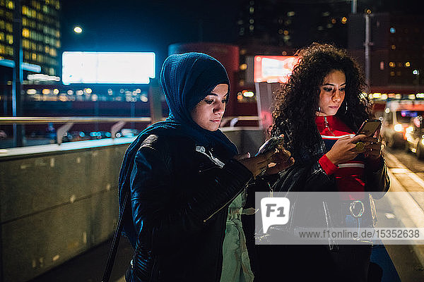 Junge Frau im Hidschab mit Freundin betrachtet nachts Smartphones in der Stadt