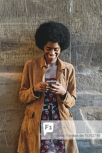 Young woman with afro hair using smartphone  leaning against stone wall