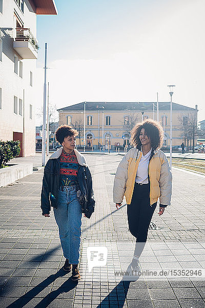 Zwei coole junge Freundinnen schlendern auf dem städtischen Bürgersteig  in voller Länge