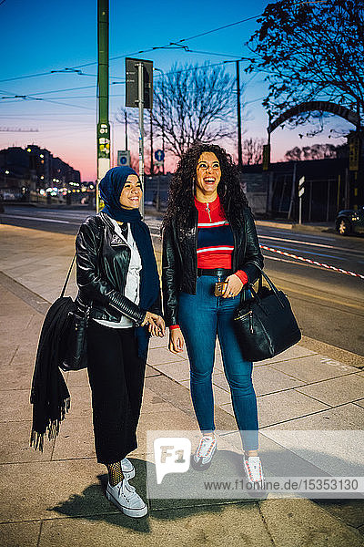 Junge Frau im Hidschab mit bester Freundin auf dem Bürgersteig der Stadt in der Dämmerung  in voller Länge