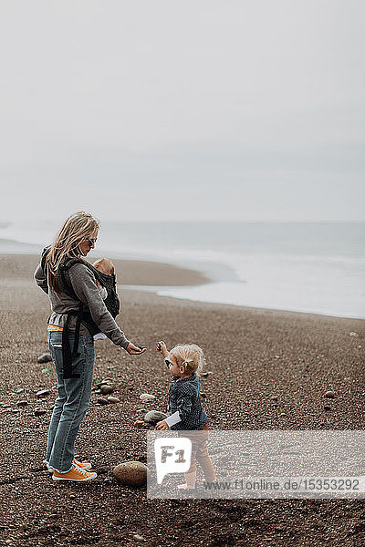 Mutter mit Baby in Tragetasche spielt mit Kleinkind am Strand